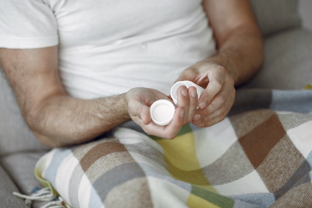 Man seated on couch with blanket on lap opening pill bottle