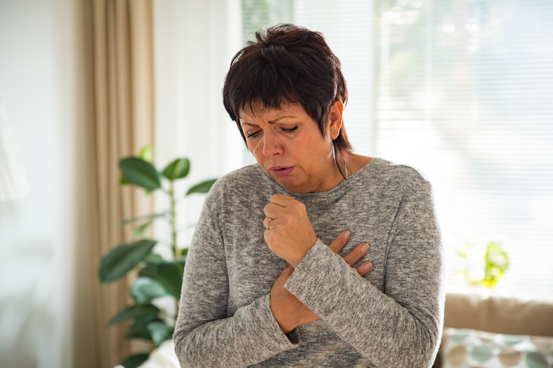 Middle-aged woman coughing in front of picture window and plant 