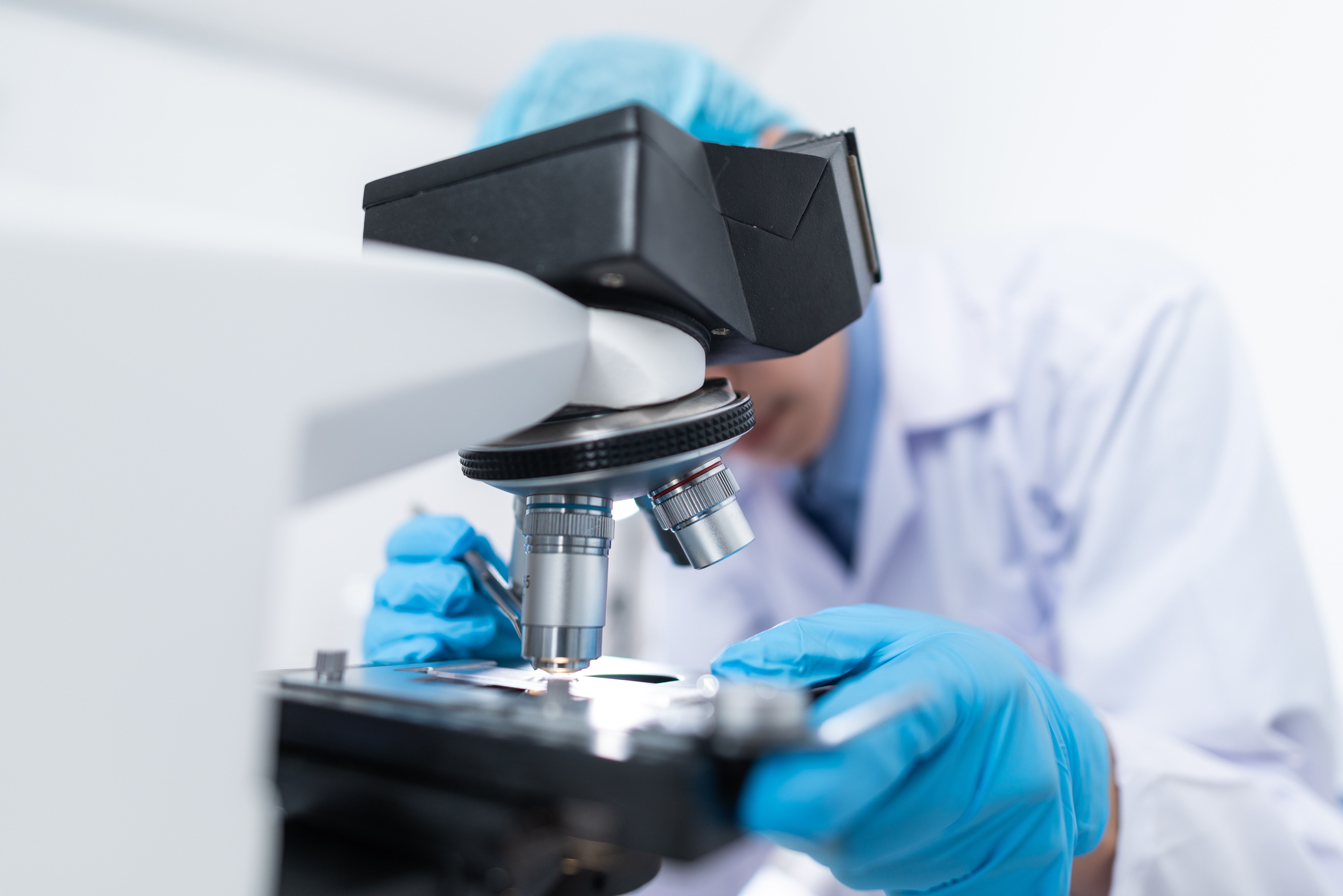 Researcher wearing gloves and cap looking into microscope