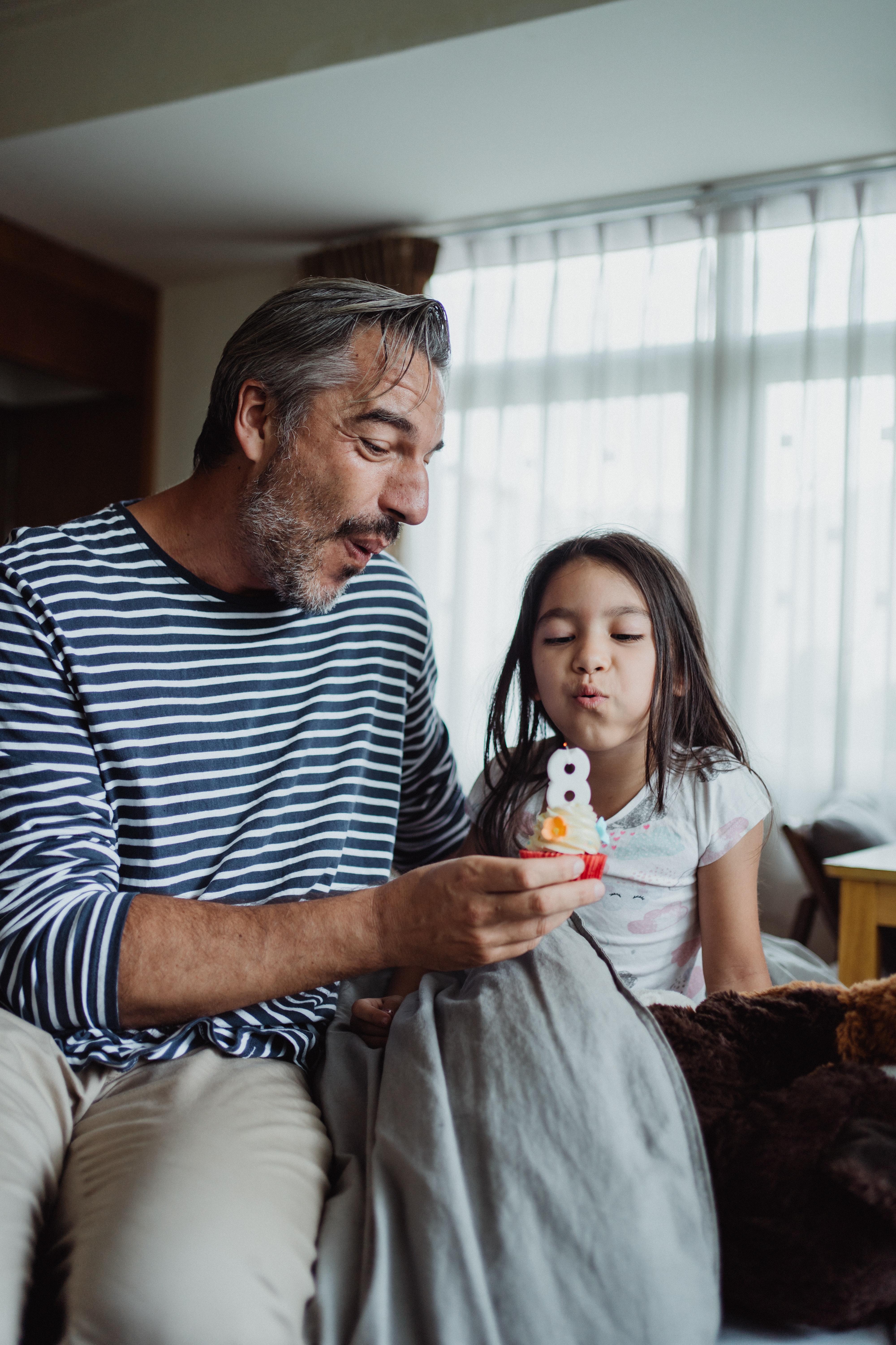 Older man sitting next to young girl blowing out a birthday candle in the shape of an 8 on a cupcake