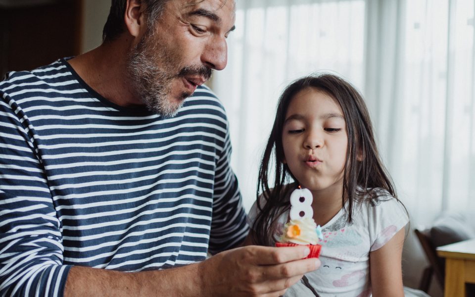 Older man sitting next to a young girl, both blowing out a number 8 candle on a cupcake